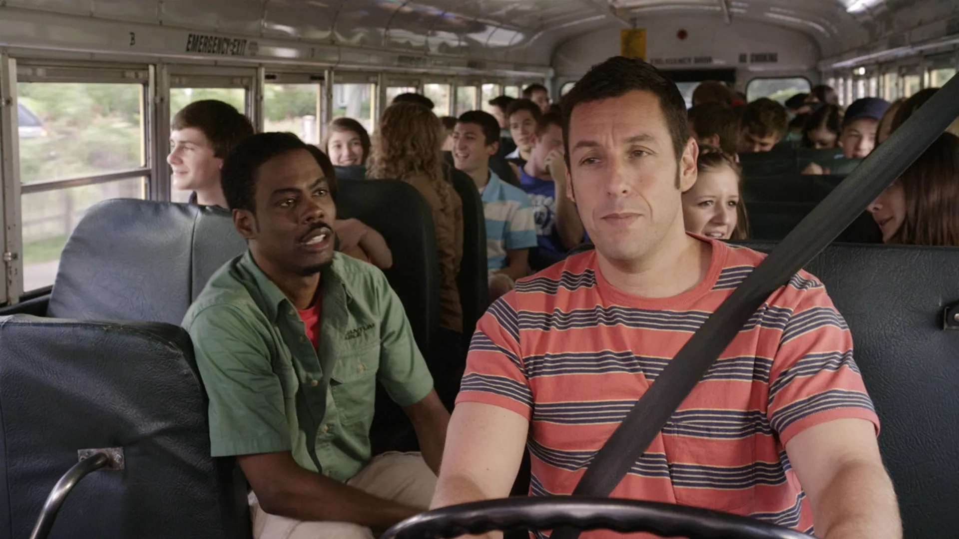 Adam Sandler driving a school bus while talking to Chris Rock in a crowded bus full of students in a movie still from Grown Ups 2.