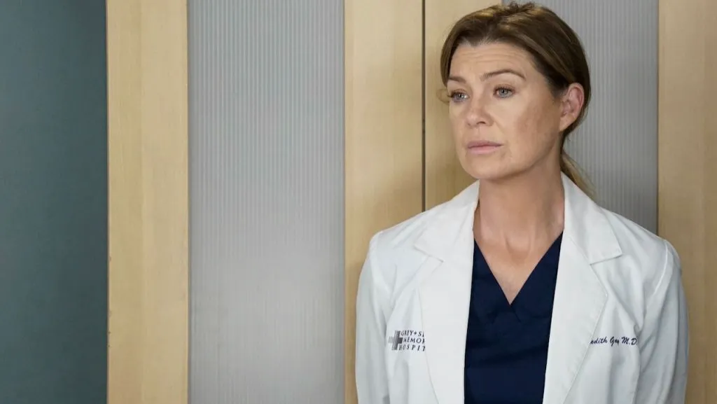 Meredith Grey, wearing her white doctor’s coat and navy scrubs, stands pensively in a hospital hallway in the TV show Grey’s Anatomy.