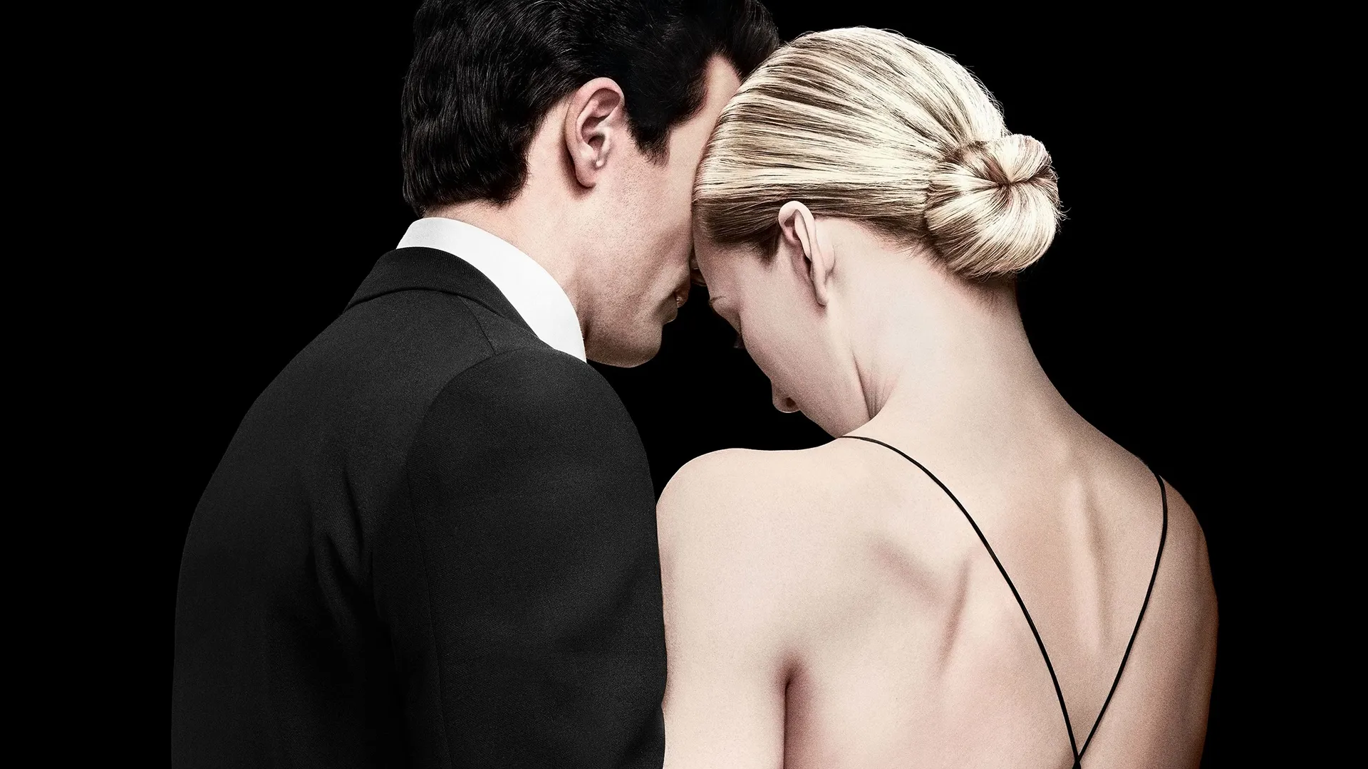 Against a solid black background, John F. Kennedy Jr. (Paul Anthony Kelly) and Carolyn Bessette (Sarah Pidgeon) stand closely with their foreheads gently touching, captured from the chest up with him in a black suit and her facing away in a low-backed gown.