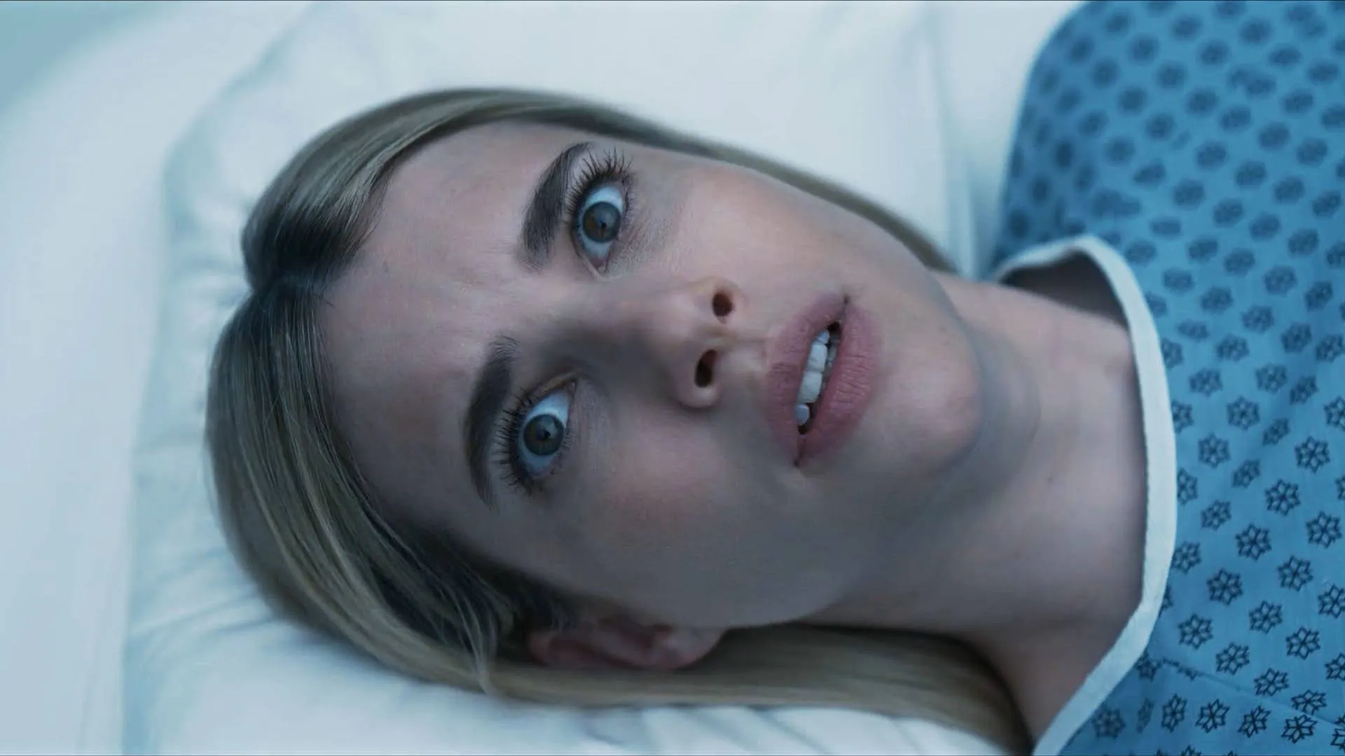 A terrified young woman, Anna Victoria Alcott (Emma Roberts), lies in a hospital bed wearing a patterned blue hospital gown, her eyes wide with shock and alarm as she stares directly upward.