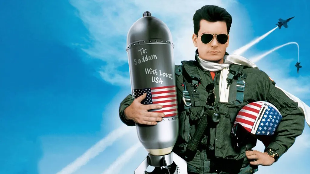 Charlie Sheen as fighter pilot Topper Harley in Hot Shots!, wearing aviator sunglasses and holding a bomb that reads “To Saddam, With Love.”