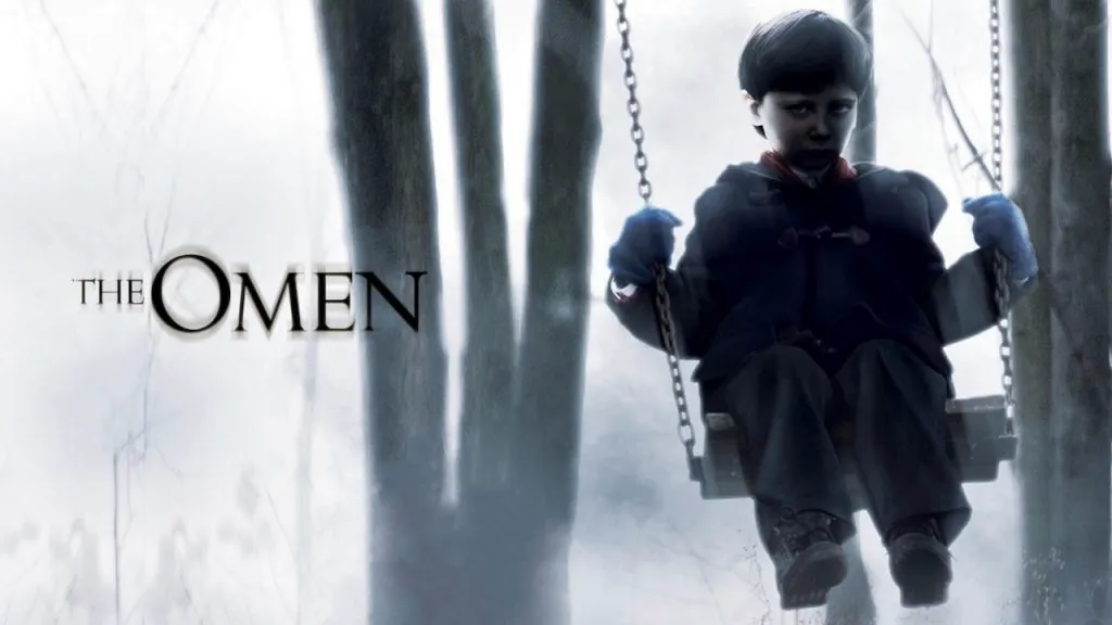 Poster for The Omen showing a pale, dark-eyed boy in a black coat sitting on a swing in a foggy forest, with the movie title displayed to the left in eerie black text.