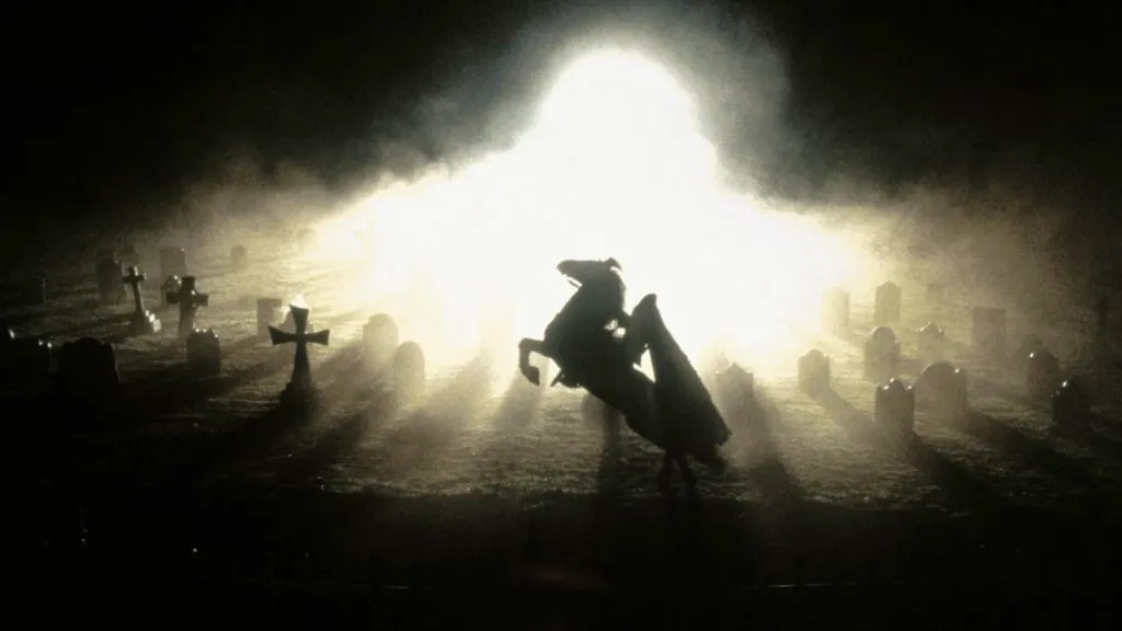 A haunting still from Sleepy Hollow showing the Headless Horseman on a rearing horse emerging from a graveyard bathed in eerie light.
