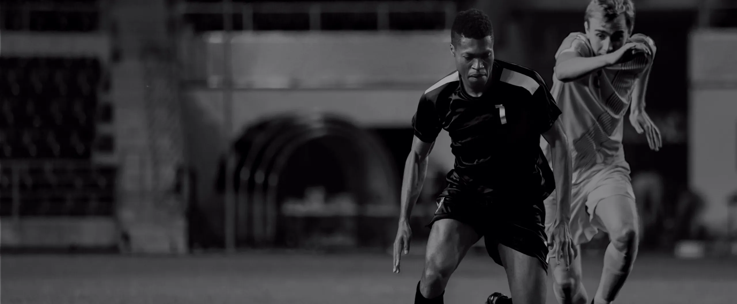 Two soccer players race down the field during a night match, the player in a dark kit breaking past a defender.