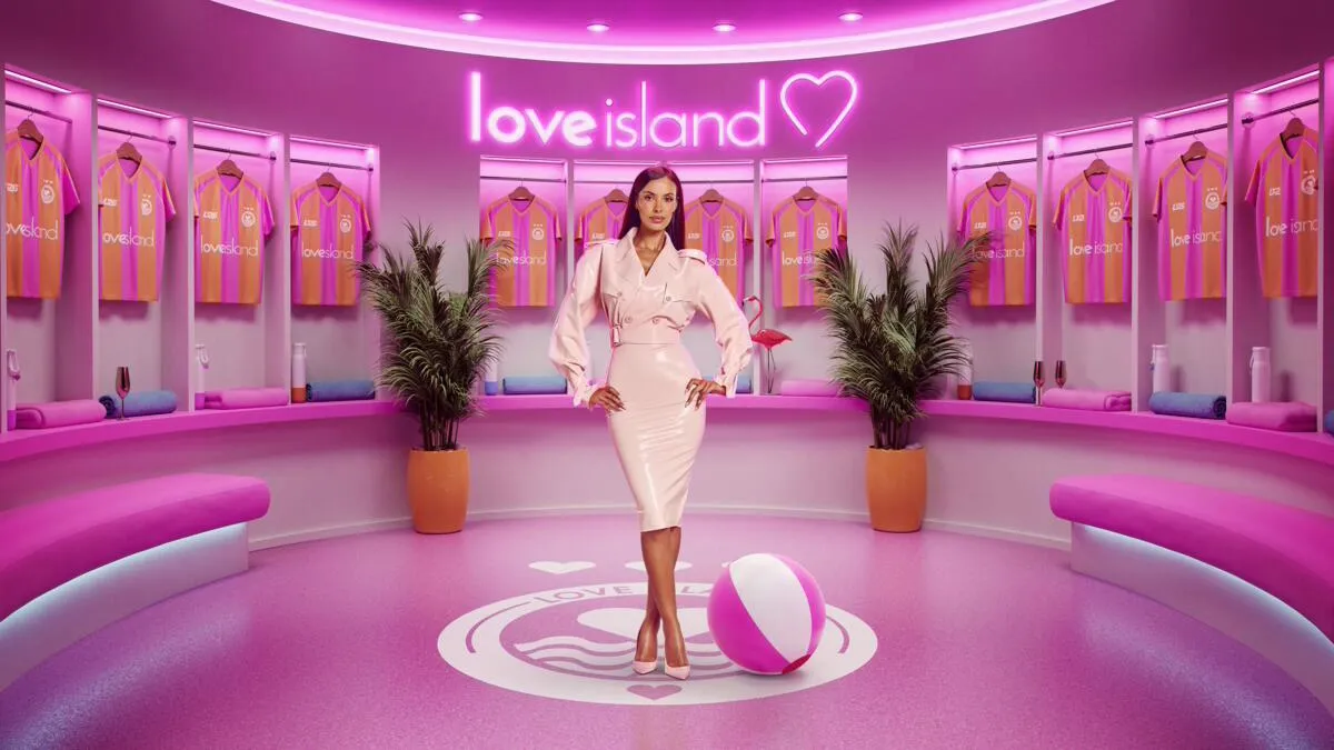 Maya Jama stands in the pink dressing room set for Love Island UK Season 13, surrounded by team jerseys and neon Love Island branding.