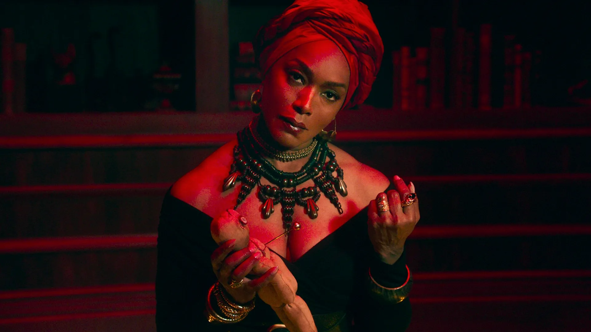 An imposing woman, Marie Laveau (Angela Bassett), looks directly forward under intense red lighting while wearing an ornate statement necklace and a large red headwrap, her hands confidently handling a doll.