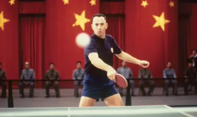 Tom Hanks as Forrest Gump playing table tennis in front of a red backdrop with yellow stars.