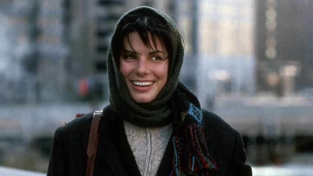 Sandra Bullock as Lucy Moderatz in While You Were Sleeping, smiling warmly while bundled up in a scarf on a cold city day.