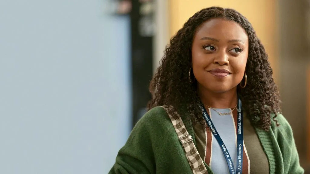 Quinta Brunson as Janine Teagues in Abbott Elementary, smiling warmly while wearing a green cardigan and school lanyard in a classroom hallway.