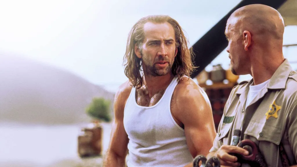 Nicolas Cage as Cameron Poe in Con Air, wearing a white tank top and long hair while talking to a guard.