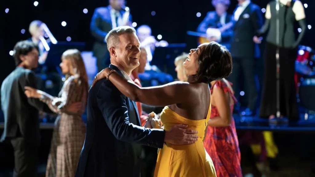 Two characters from the TV show 9-1-1 smiling and dancing together at an elegant gala with a live band performing in the background.