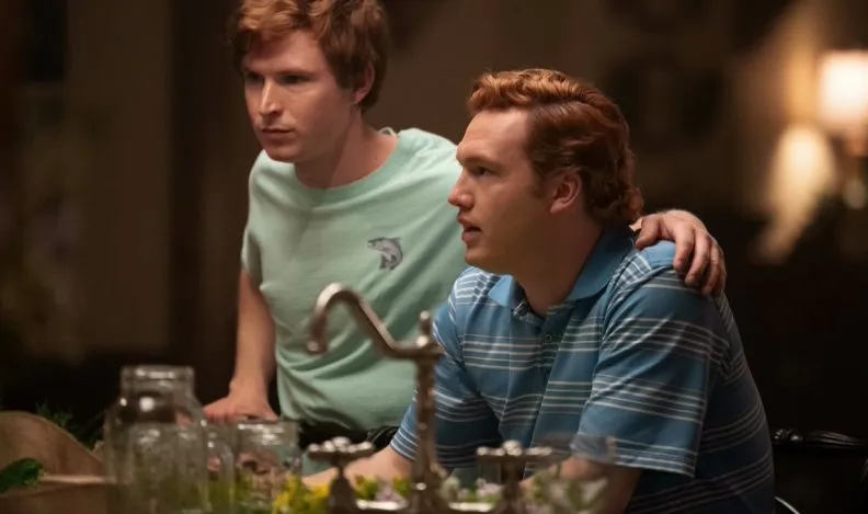 Two men portraying members of the Murdaugh family sit together at a dining table, one resting his hand on the other’s shoulder in a warmly lit room.
