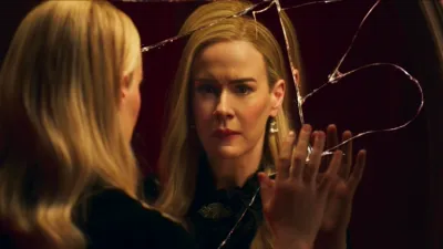 An elegant blonde woman, Cordelia Goode (Sarah Paulson), stares with a distressed and emotional expression at her reflection in a severely shattered mirror, her hand pressed flat against the cracked glass.