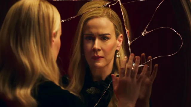 An elegant blonde woman, Cordelia Goode (Sarah Paulson), stares with a distressed and emotional expression at her reflection in a severely shattered mirror, her hand pressed flat against the cracked glass.